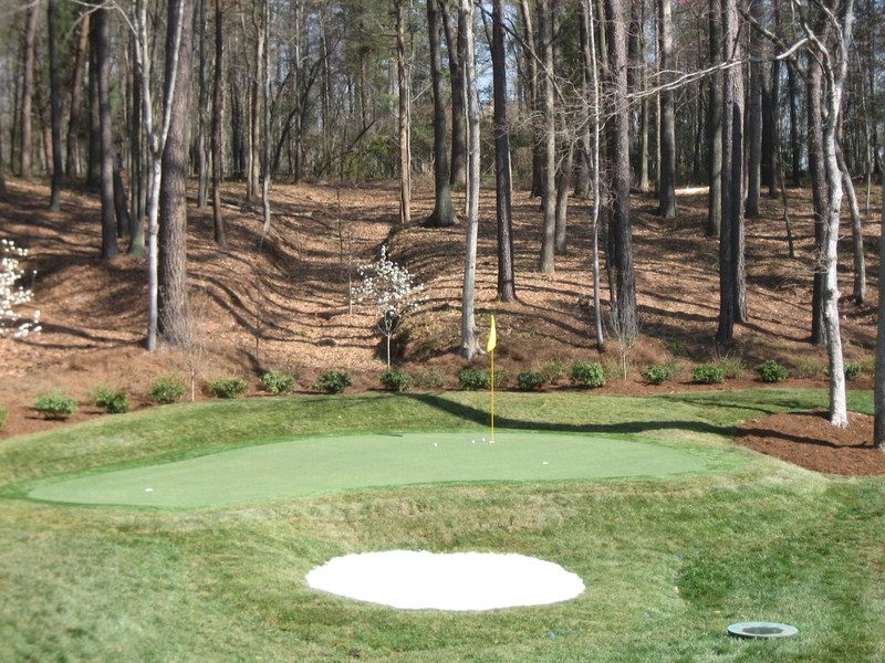 Synthetic golf green with sand bunker set against trees